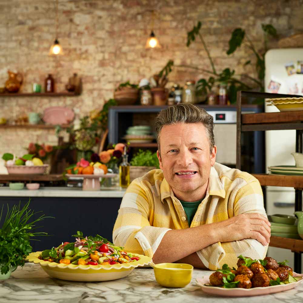 Jamie Oliver sitting at table with serving platters full of food - Robins Kitchen